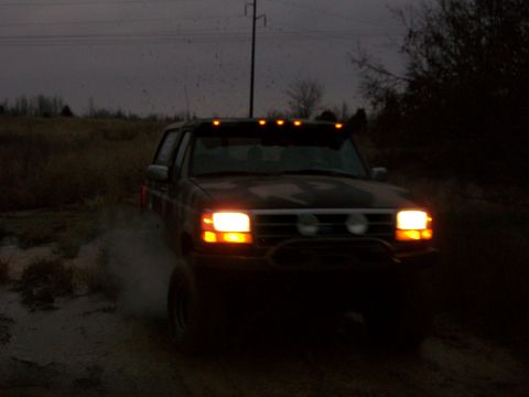 1995 Ford Bronco - 1995 Ford Bronco navigating a muddy terrain at dusk, headlights illuminated.