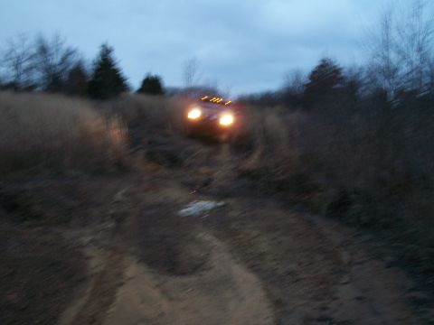 1995 Ford Bronco - A red SUV navigates a muddy dirt road as dusk settles, its headlights illuminating the path ahead.