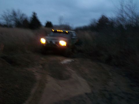 1995 Ford Bronco - This dark SUV navigates an off-road path at dusk, its headlights illuminating the muddy terrain.