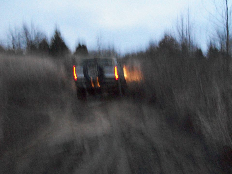 SUV driving on a dirt road with trees in the background, taillights are on