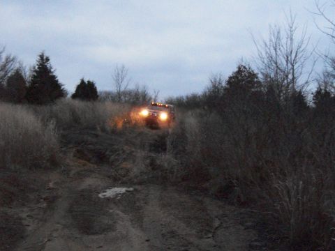 1995 Ford Bronco - A red Ford Bronco navigates a muddy trail surrounded by dry vegetation and trees under an overcast sky.