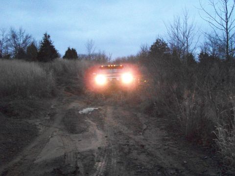 1995 Ford Bronco - A vehicle navigates a muddy path at dusk, its headlights cutting through the dim light.