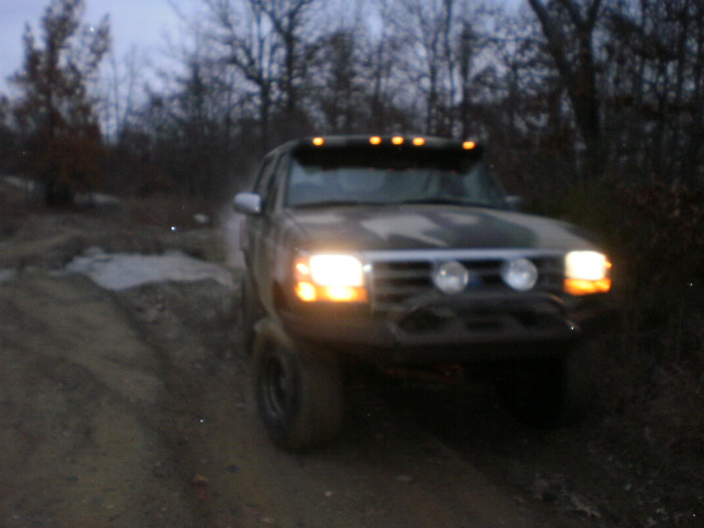 1995 Ford Bronco driving on a dirt road at dusk, headlights on