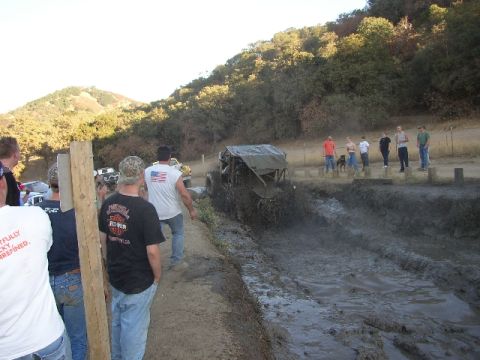 1989 Ford Bronco - An off-road vehicle navigates a muddy track as onlookers watch from the side.