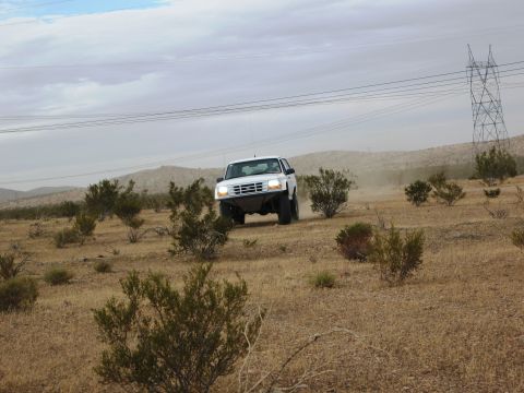 1986 Ford Bronco - A white 1992 Ford Bronco navigates a dusty, off-road terrain under a cloudy sky.