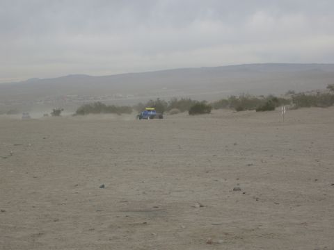 1986 Ford Bronco - A blue off-road buggy navigates a dusty desert terrain, with another vehicle visible in the distance.