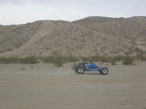 1986 Ford Bronco - A blue dune buggy kicks up dust as it drives through a desert landscape under a cloudy sky.