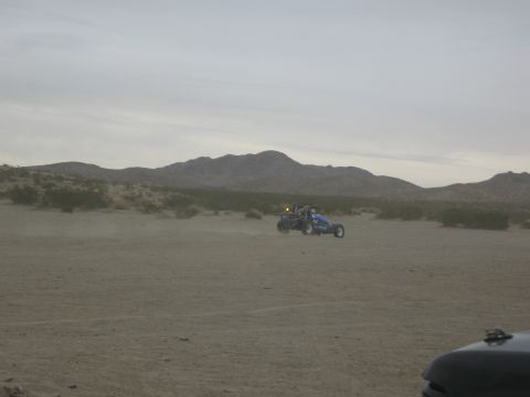 1986 Ford Bronco - A blue dune buggy kicks up dust as it drives across a desert landscape under a cloudy sky.
