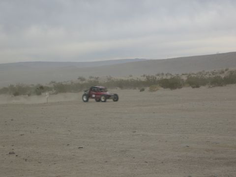 1986 Ford Bronco - A red off-road buggy kicks up dust as it drives across a sandy desert terrain under an overcast sky.
