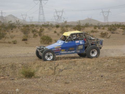 1986 Ford Bronco - An off-road buggy with a roll cage and racing decals navigates a dusty desert terrain under a cloudy sky.