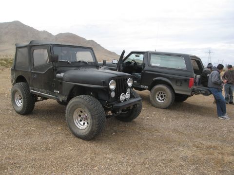 1986 Ford Bronco - A black Jeep and a black Ford Bronco are parked side-by-side on a gravelly surface with mountains in the background.