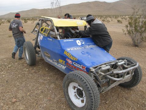 1986 Ford Bronco - An off-road buggy with a roll cage and large tires is being worked on by people in a desert environment.
