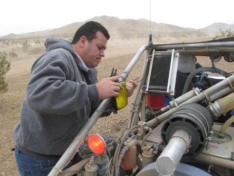 1986 Ford Bronco - Man performs maintenance on a custom off-road vehicle's engine in a dusty, desert environment.