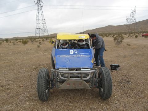 1986 Ford Bronco - A desert buggy with racing occupants and a mechanic stands ready under power lines.