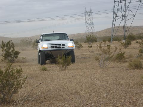 1986 Ford Bronco - A white off-road truck navigates a desert landscape under a cloudy sky, with power line towers visible.