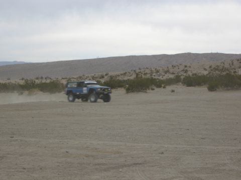 1986 Ford Bronco - A blue SUV kicks up dust as it drives through a desert landscape.