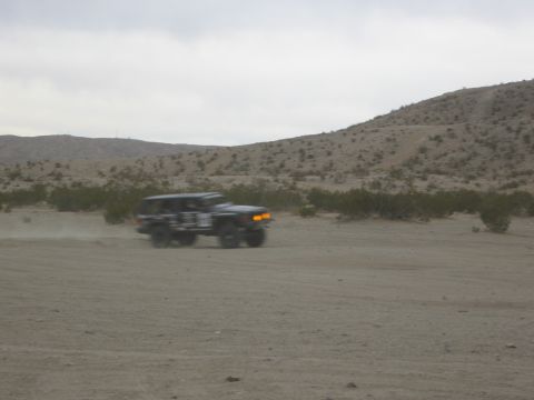 1986 Ford Bronco - An SUV kicks up dust as it drives through a dry, desert terrain under a cloudy sky.