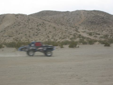 1986 Ford Bronco - A modified off-road truck kicks up dust as it drives through a desert environment under a cloudy sky.