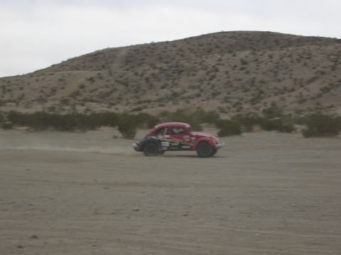 1986 Ford Bronco - A red custom beetle-style car with racing decals is seen driving on a dirt road in a desert landscape.