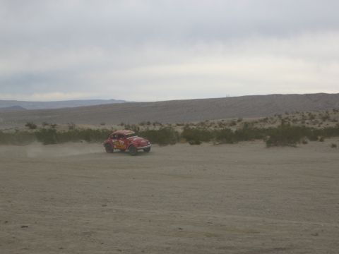 1986 Ford Bronco - A red Volkswagen Beetle races across a dusty desert landscape under an overcast sky.