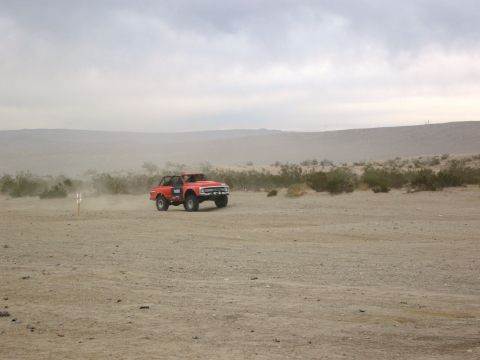 1986 Ford Bronco - A red off-road vehicle kicks up dust as it drives through a barren, desert environment under a cloudy sky.