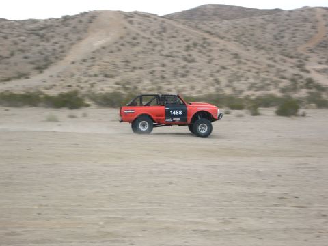 1986 Ford Bronco - An orange off-road vehicle with a roll cage drives through a dusty, desert landscape.