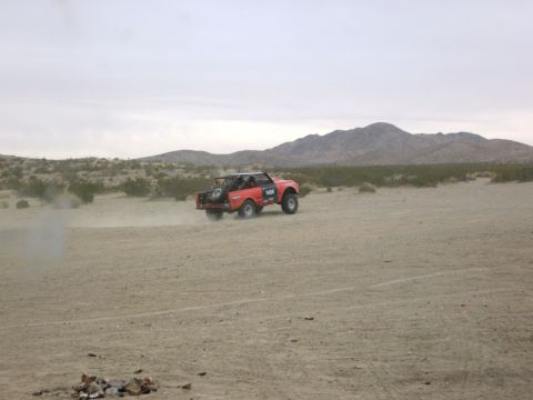 1986 Ford Bronco - A red off-road vehicle kicks up dust as it drives through a desert landscape under a cloudy sky.