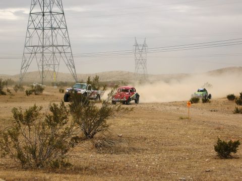 1986 Ford Bronco - Multiple off-road vehicles compete in a dusty desert race under a cloudy sky, with large power line towers in the background.