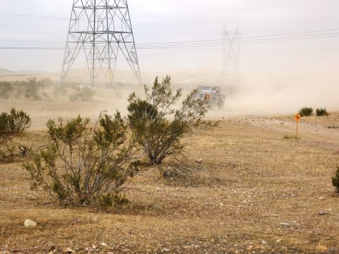 1986 Ford Bronco - An off-road vehicle navigates a dusty desert track under a hazy sky, with large power line towers in the background.