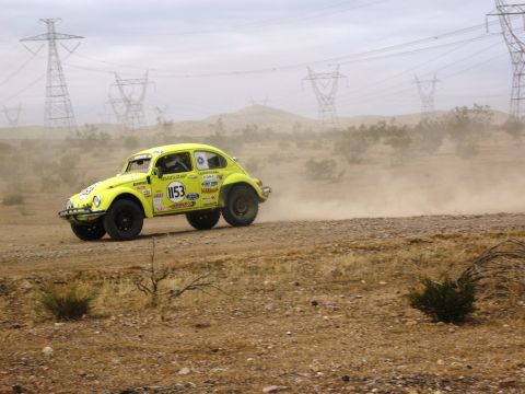1986 Ford Bronco - A yellow Volkswagen Beetle, modified for off-road racing, speeds down a dusty track with power lines visible in the distance.