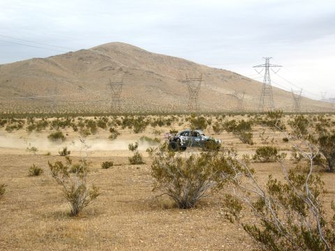 1986 Ford Bronco - A race truck navigates a dusty desert terrain with power lines and mountains in the background.