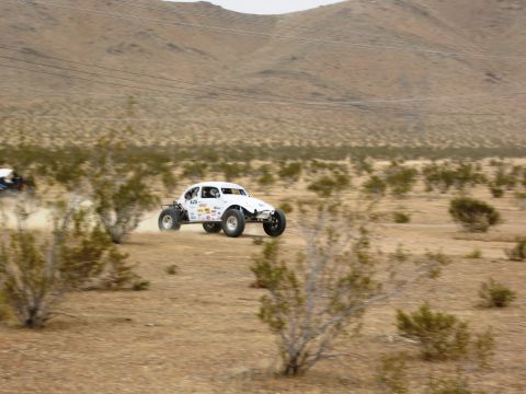 1986 Ford Bronco - A white off-road buggy races across a dusty desert landscape, with another vehicle visible in the distance.