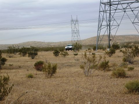 1986 Ford Bronco - A white SUV drives through a dry, brush-filled desert landscape under a cloudy sky, with large power line towers in the foreground.