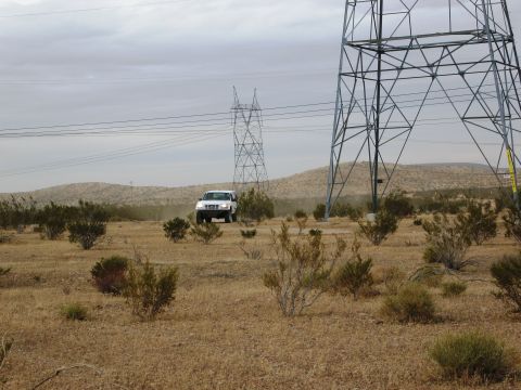 1986 Ford Bronco - A white SUV navigates a dusty desert terrain under a cloudy sky, with large power line towers nearby.
