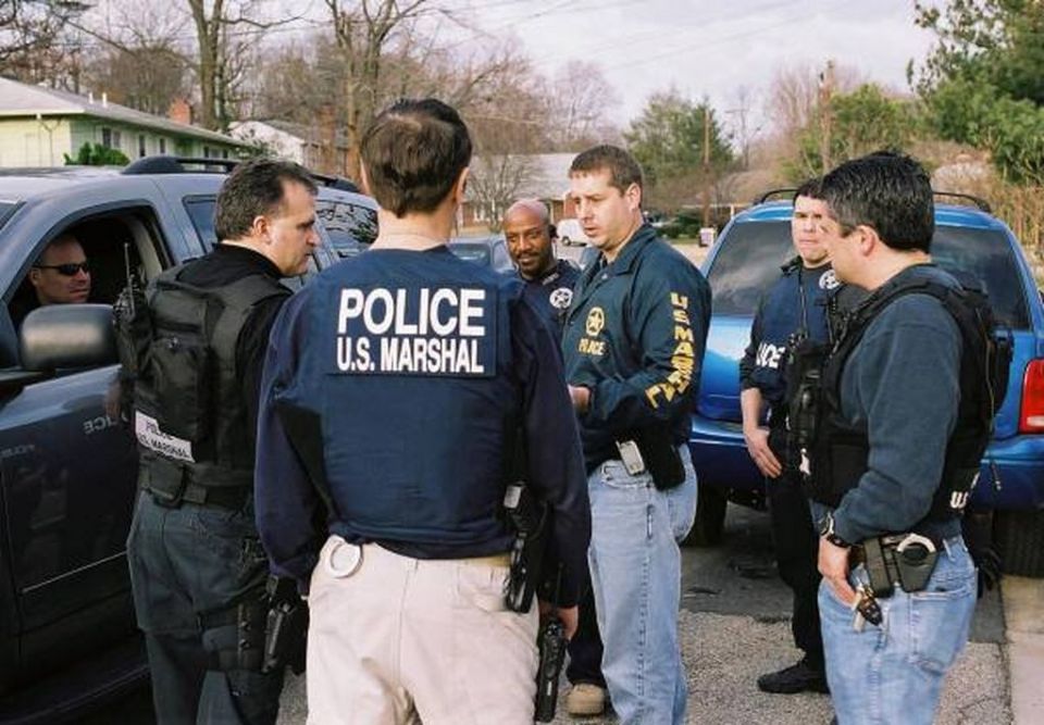 Several police officers in tactical gear are gathered around two dark-colored SUVs