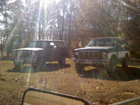 1990 Ford Bronco - Two Ford Broncos are parked outdoors on a sunny day with trees in the background.