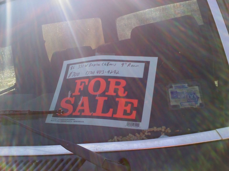 Rear view of a dark-colored Ford Bronco with a "FOR SALE" sign on the windshield, showing a 1986 engine detail