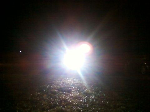 1990 Ford Bronco - A powerful headlight illuminates the ground at night, with a faint hint of a vehicle on the right.