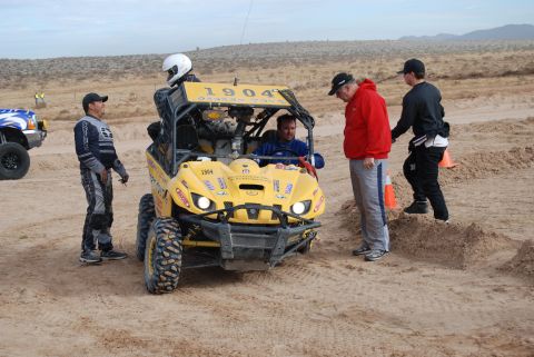 1982 Ford Bronco - People interact around a yellow off-road buggy in a sandy, desert environment.