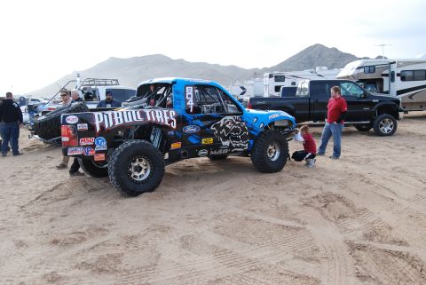 1982 Ford Bronco - A custom off-road truck with racing decals is shown in a sandy area with other vehicles and RVs in the background.