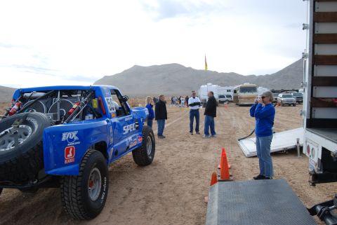 1982 Ford Bronco - A blue off-road racing truck with a spare tire mounted is parked in a desert area with spectators and RVs in the background.