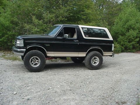 1993 Ford Bronco - Side profile of a black and tan two-tone Ford Bronco, lifted with off-road tires and alloy wheels, parked on gravel near trees.