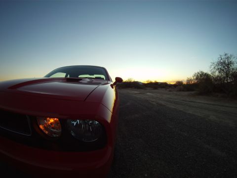 2010 Dodge Challenger - A red Dodge Challenger is captured in a low-angle shot at sunset, parked on a gravel road with desert scrub in the background.