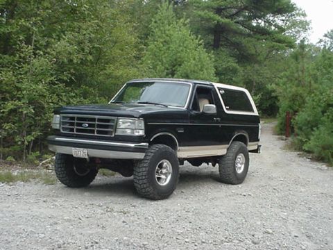 1993 Ford Bronco - Black Ford Bronco with a white removable hardtop and tan rocker stripe, parked on a gravel trail at the edge of the woods and shot from the…