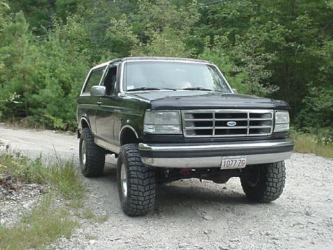 1993 Ford Bronco - Three-quarter front view of a lifted dark green full-size Ford Bronco with a white removable hardtop, parked on a gravel road against a wal…
