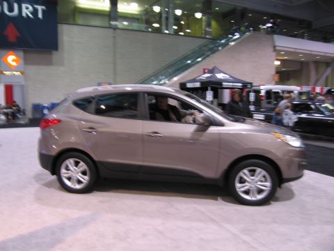 1991 Ford LTD Crown Victoria - A tan Hyundai Tucson is displayed indoors, with people and other vehicles visible in the background.