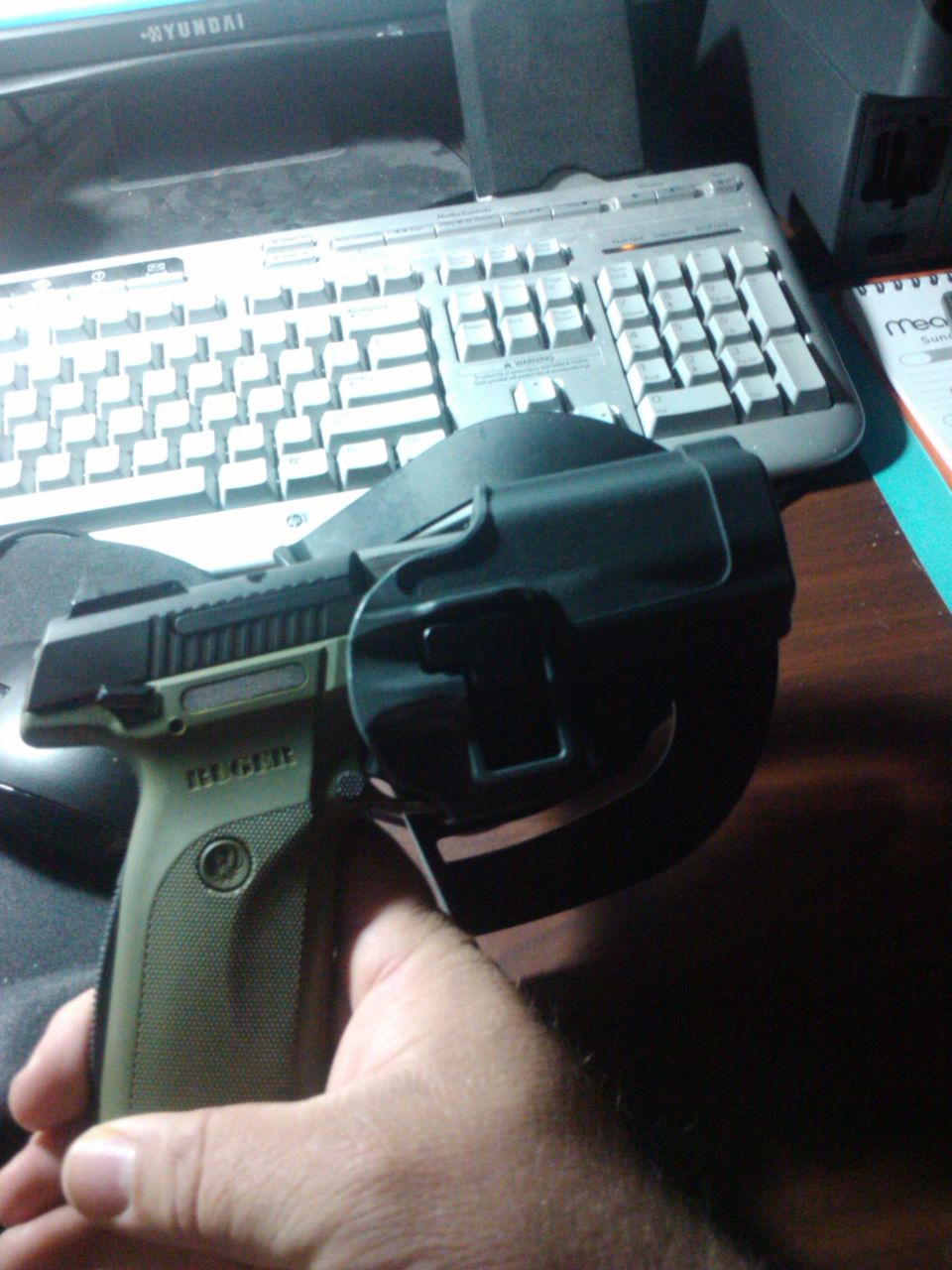 Hand holding a tan and black pistol in a black holster, with a keyboard and monitor in the background