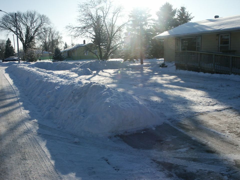 Snow piled high on a residential street and driveway with a house in the background