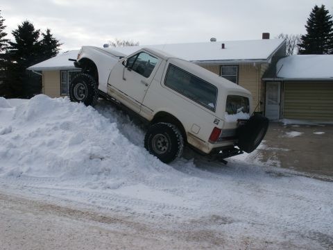 1966 Ford Bronco - A white SUV navigates a steep snowdrift, its tires digging into the snow for traction.