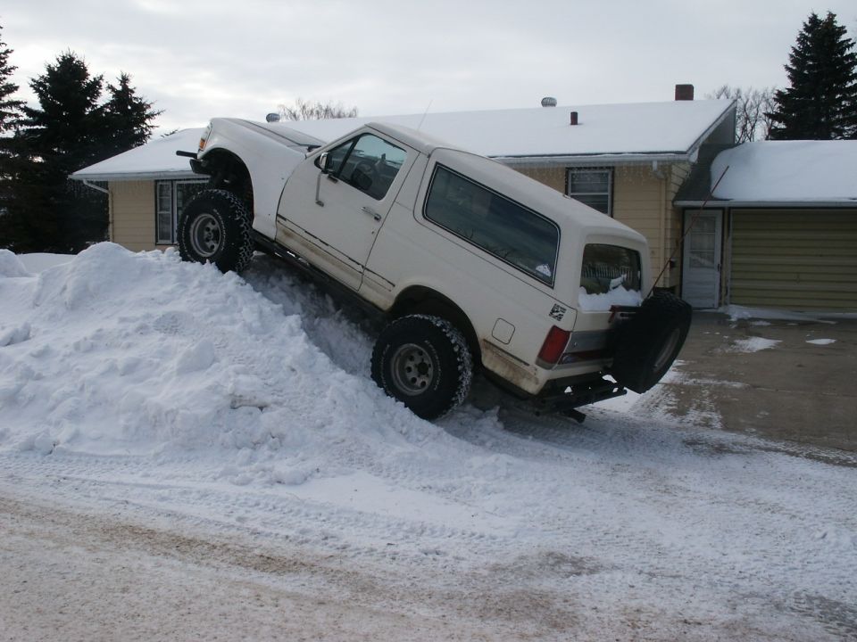 White SUV with lifted suspension and off-road tires climbing a large snow pile in front of a house